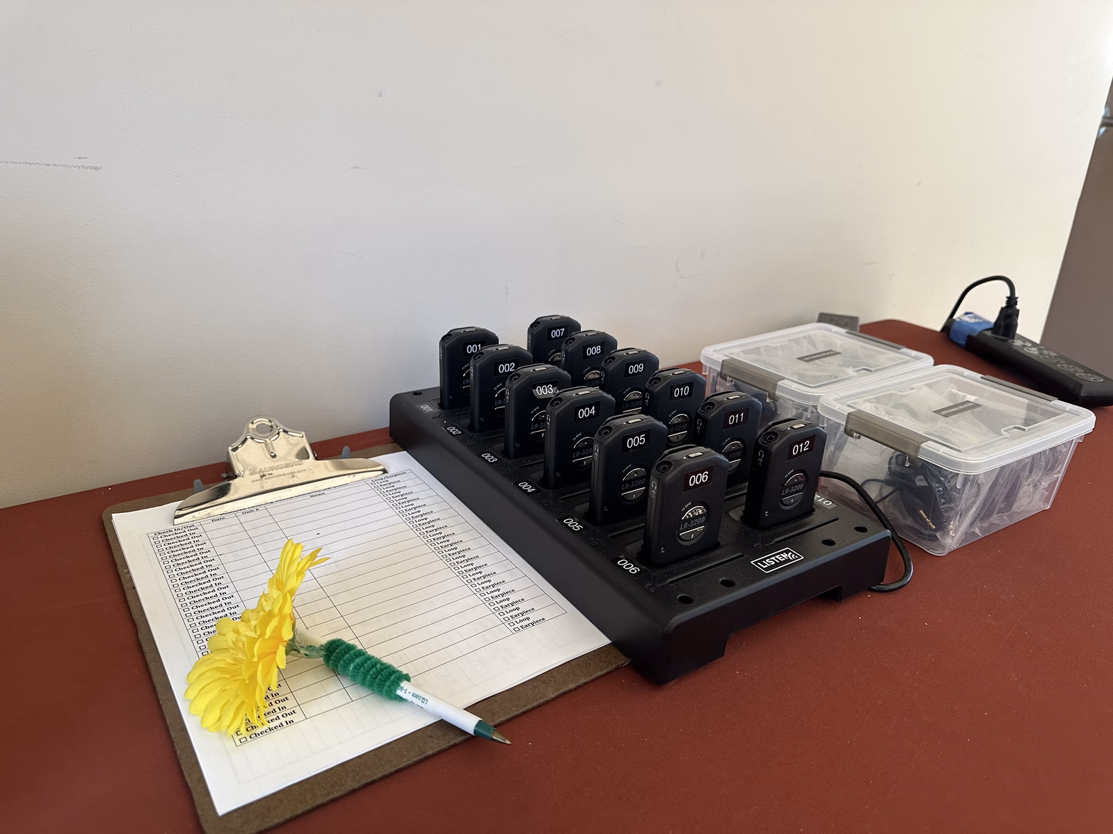 A charging rack of numbered assisted listening receivers beside a clipboard sign-out sheet and bins of earpieces