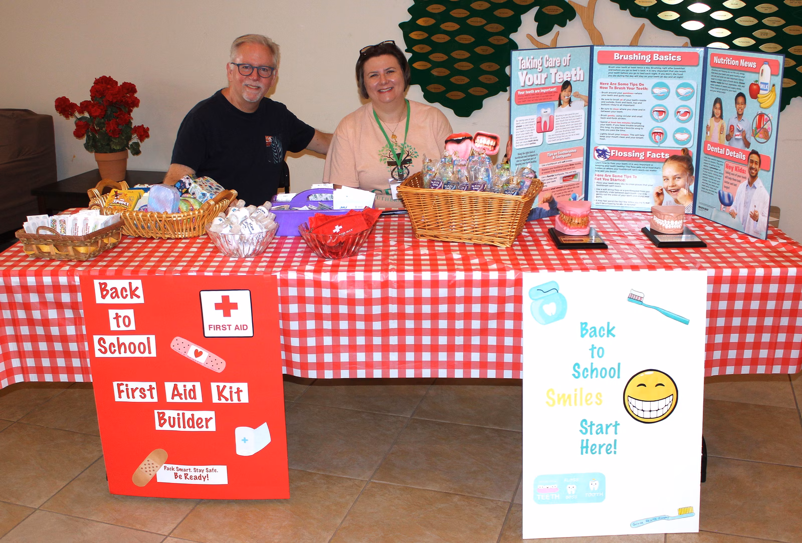 Two volunteers seated at a health ministry table with baskets of first aid and dental supplies, a Back to School First Aid Kit Builder sign, and posters on brushing and nutrition.