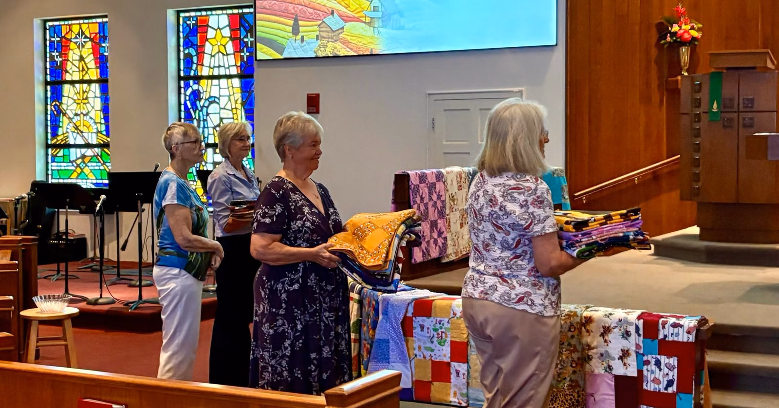Four women standing at the front of the sanctuary during the blessing of the quilts, holding folded handmade quilts with more draped over the chancel rail behind them.