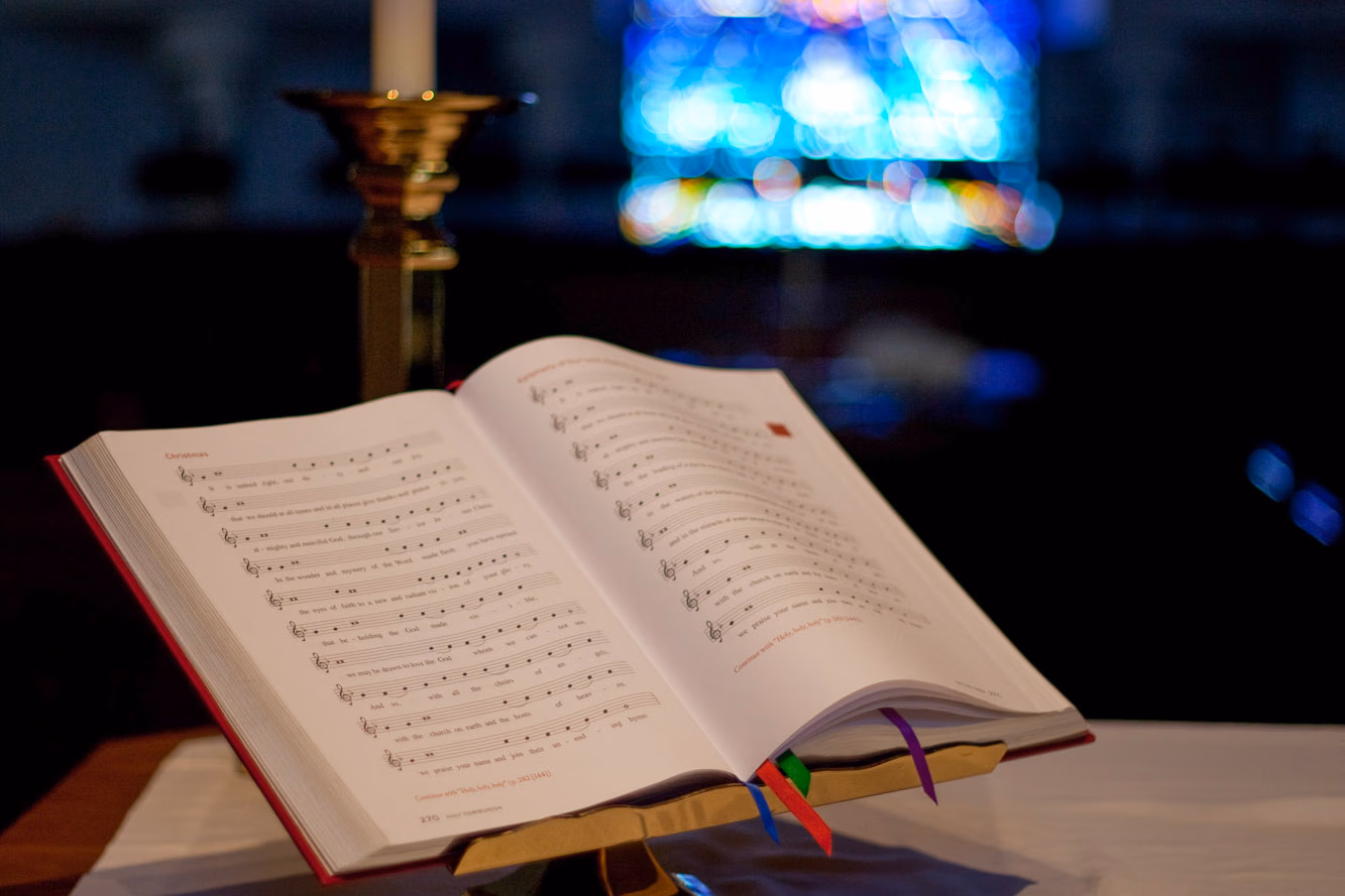 An open liturgical worship book on a brass stand at the altar, lit by a candle with a stained glass window behind