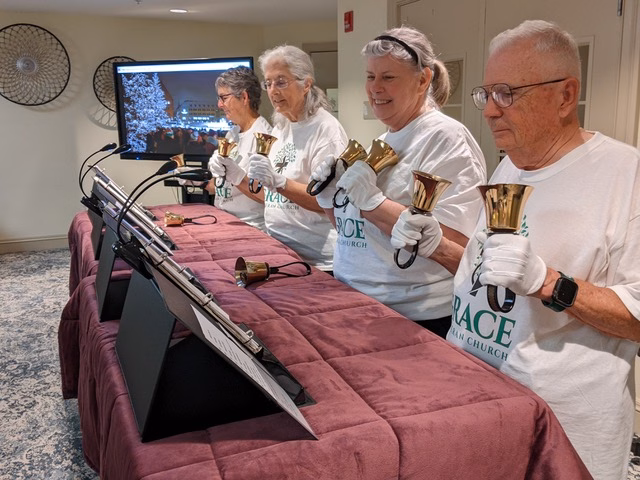 Four handbell ringers in white Grace Lutheran T-shirts and white gloves standing behind a draped table, lifting polished handbells as they play a mini-concert in a community room.