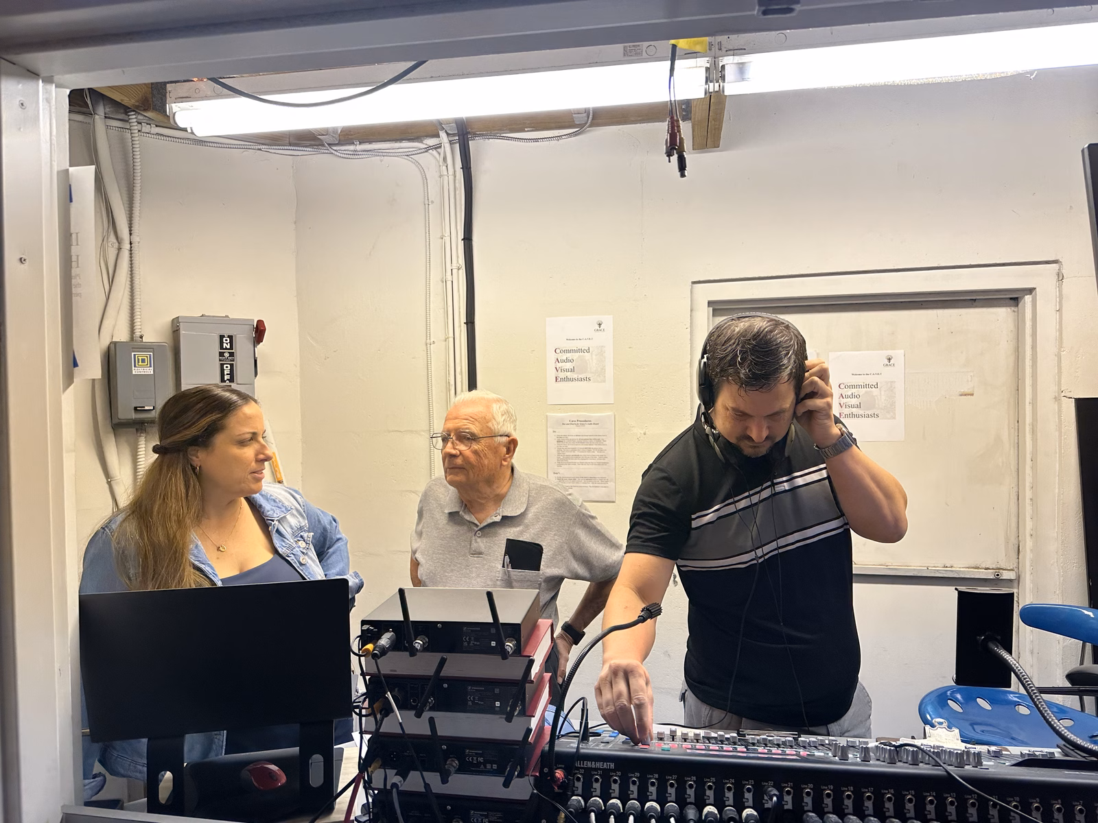 Three CAVE volunteers in the church's audio booth: a man wearing headphones adjusts a sound mixing board while a woman and an older man look on beside a stack of wireless microphone receivers, with Committed Audio Visual Enthusiasts signs posted on the wall behind them.