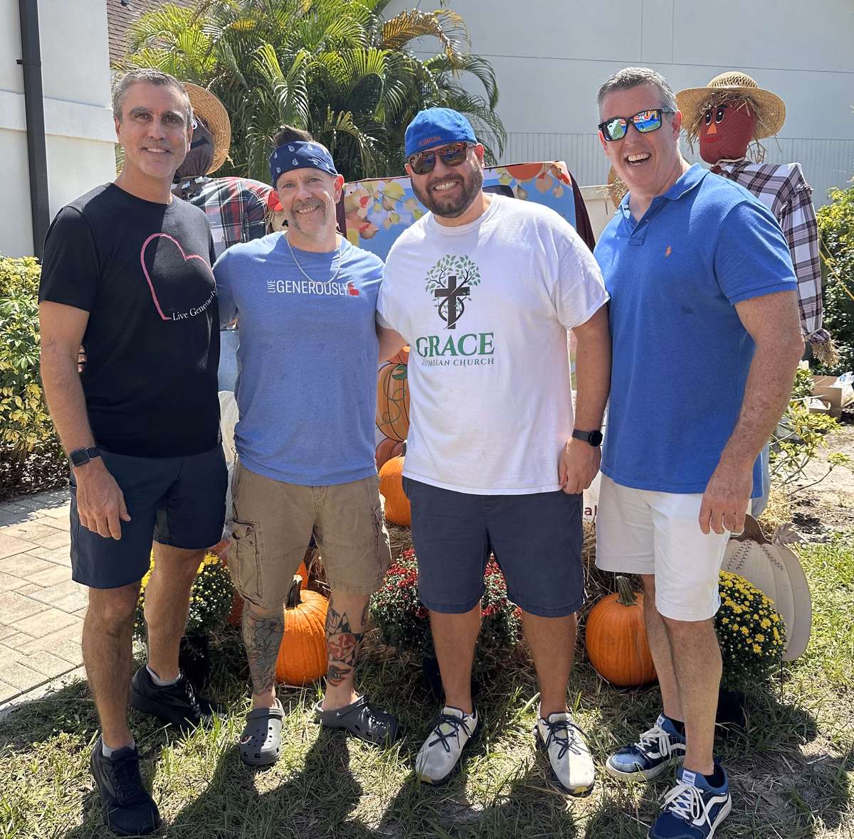 Four men standing arm in arm on the church lawn on a sunny day, smiling at the camera in front of a fall display of scarecrows, pumpkins, and potted mums; one wears a Grace Lutheran Church T-shirt and two wear Live Generously shirts.