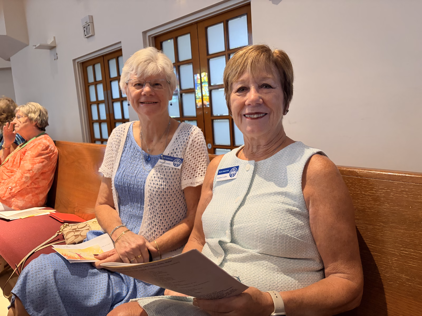Two smiling women sit side by side in a wooden pew holding worship bulletins, wearing name tags, with other worshippers seated nearby before a service.