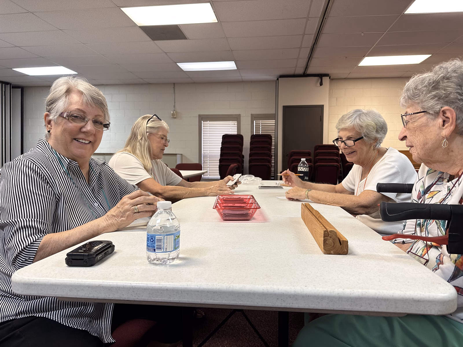 Four women seated around a long table in Holmer Hall playing cards together, hands of cards held low and a card tray between them, smiling and talking.