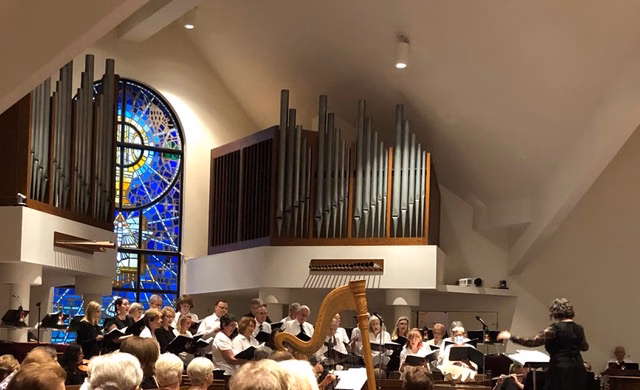 The Grace choir singing at the front of the sanctuary during the Lessons and Carols service, beneath the tall organ pipes and the blue stained glass window, with a harpist and chamber musicians alongside them and the director conducting at right.