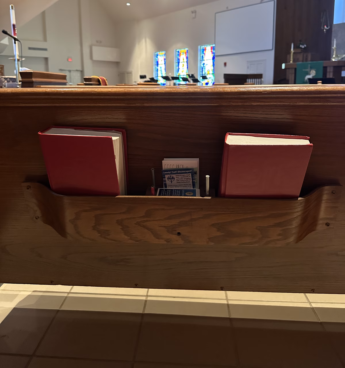 The back of a wooden pew with two red hymnals in its rack and a center holder stocked with pencils and Stephen Ministry cards, the sanctuary's stained glass windows visible beyond.