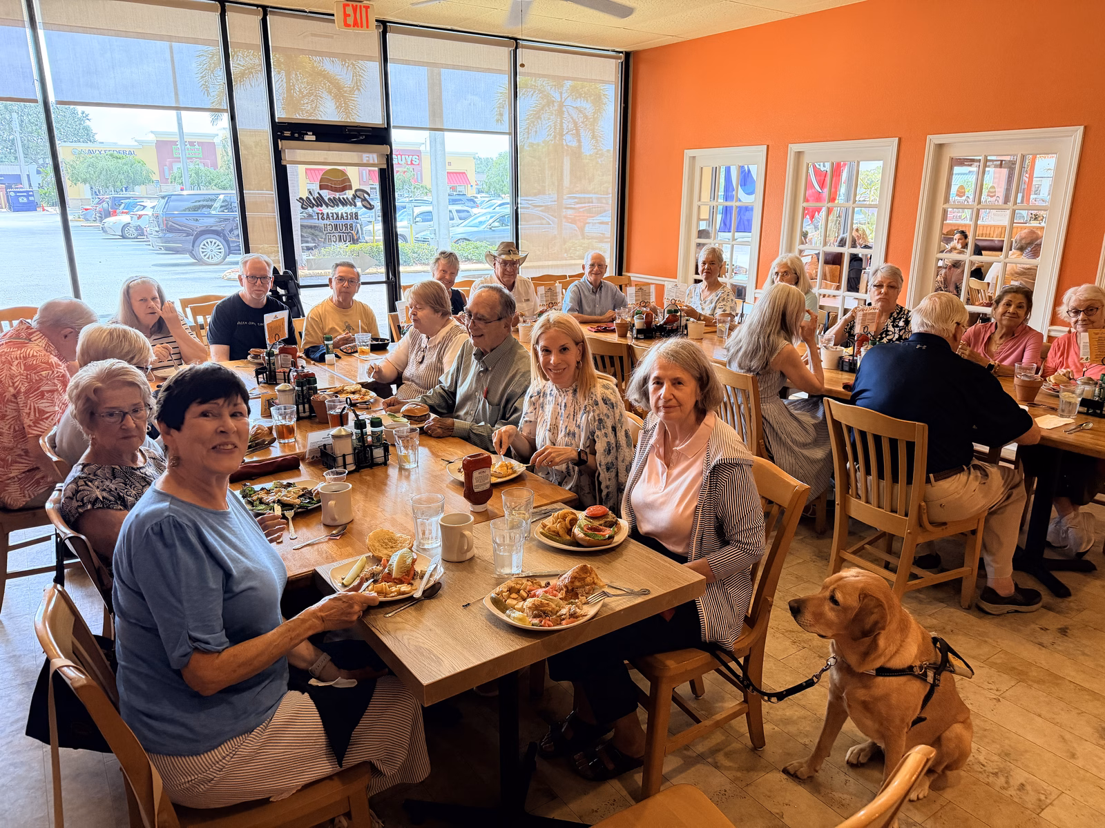 A large group of church members seated together at long joined tables in a bright restaurant, plates of breakfast and lunch in front of them, many turned toward the camera; a yellow service dog in a harness sits beside the table at right.