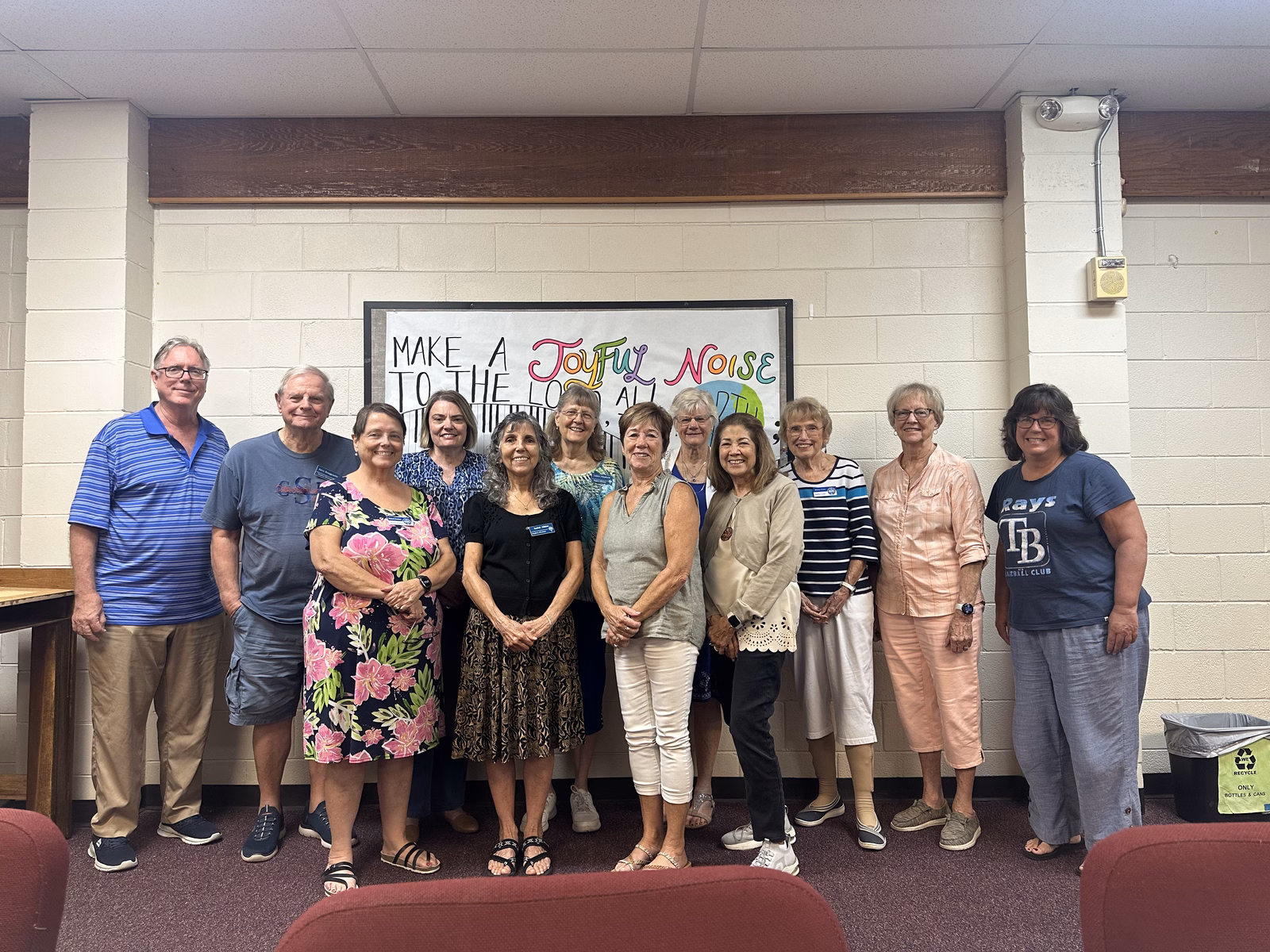 Twelve Stephen Ministers standing together in a row in Holmer Hall, smiling toward the camera in front of a hand-lettered Make a Joyful Noise banner.