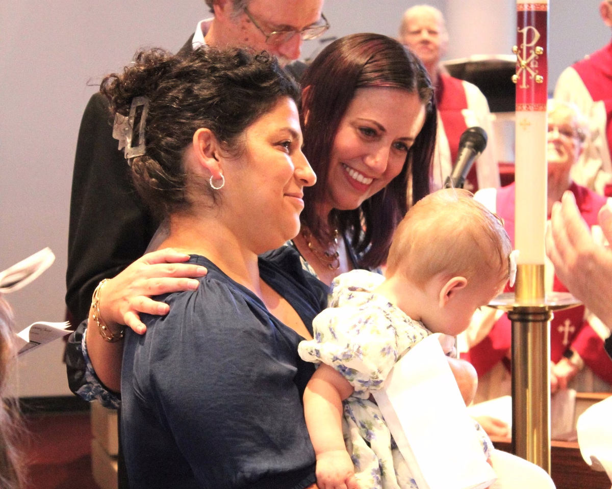 A baby held close in a family member's arms during a baptism, with the tall Paschal candle beside them and family and choir gathered around