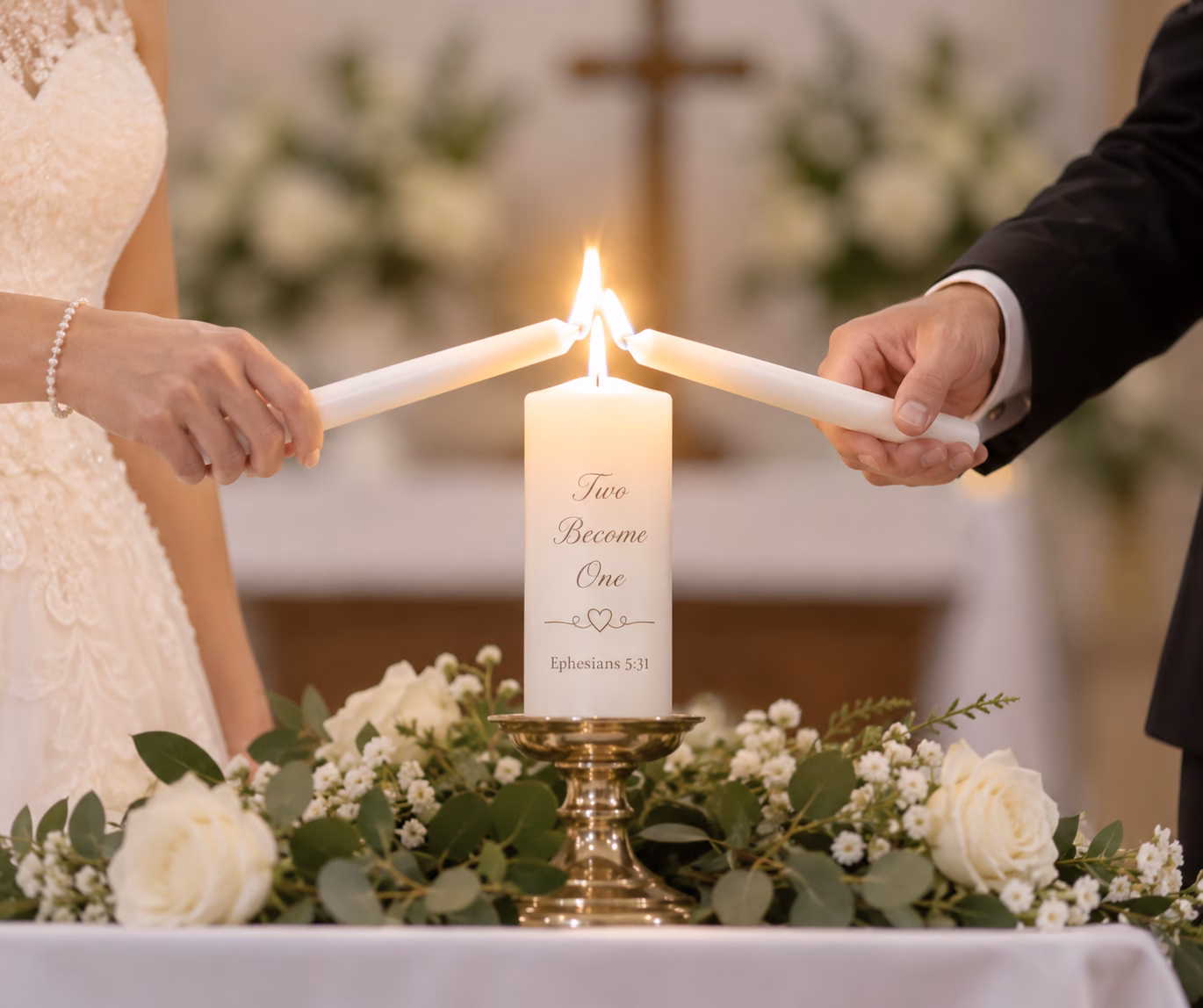 A bride and groom each holding a lit taper to a white unity candle inscribed "Two Become One" and Ephesians 5:31, set in a brass holder among white roses and greenery, with a cross and altar flowers blurred behind